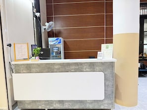 A reception desk with a wooden panel wall, a fan, and a blue refrigerator. - Doi Ngoai Bien Phong SPA Hotel (Buon Ma Thuot)