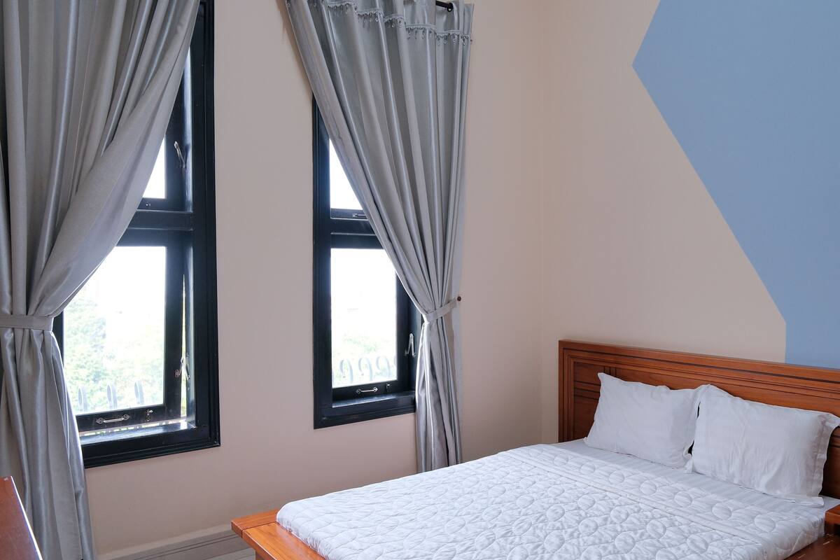 A bedroom with a wooden headboard, a white quilted bedspread, and two windows with curtains.