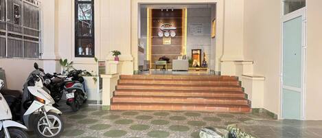 A hotel lobby with a reception desk, a staircase leading to the upper floor, and several parked scooters.