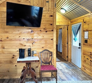 A wooden interior with a flat-screen TV, a table with coffee makers, a chair, and a door with a curtain.