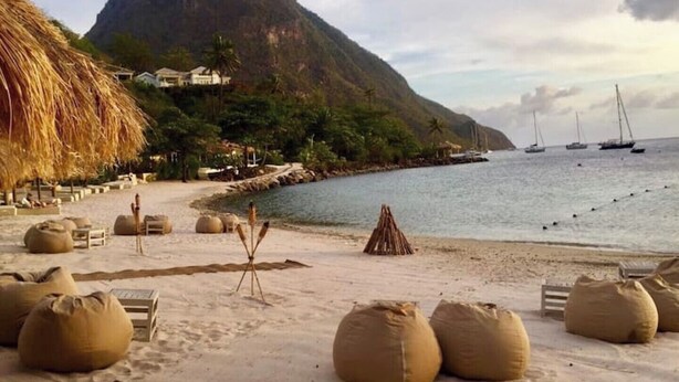 A beach with thatched-roof huts, large sandbags, and sailboats in the water. - 2-bedroom villa with WiFi, AC in St. Lucia-Beach, Falls,Pitons, Mud Bath (Laborie)