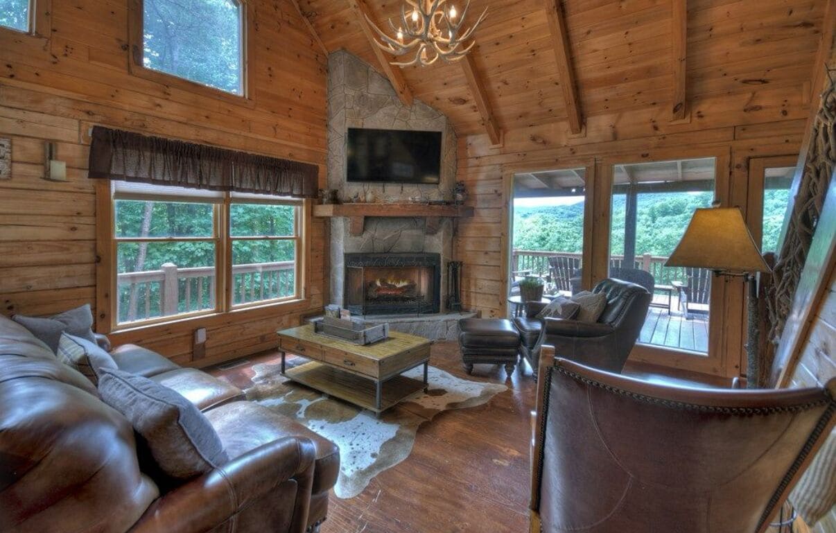 A cozy cabin interior with a stone fireplace, leather furniture, and large windows offering a view of the forest.