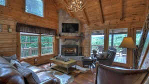 A cozy cabin interior with a stone fireplace, leather furniture, and large windows offering a view of the forest. - Luxury Pet-Friendly Mountain Cabin - Hot Tub - Views - 20 Min to Blue Ridge (Cherry Log)