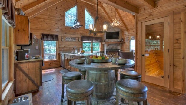 A rustic cabin interior with a wooden interior, a central island with stools, a fireplace, and a television mounted on the wall. - Luxury Pet-Friendly Mountain Cabin - Hot Tub - Views - 20 Min to Blue Ridge (Cherry Log)