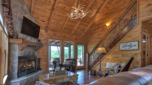 A cozy log cabin interior with a stone fireplace, leather sofa, and a staircase leading to an upper level. - Luxury Pet-Friendly Mountain Cabin - Hot Tub - Views - 20 Min to Blue Ridge (Cherry Log)