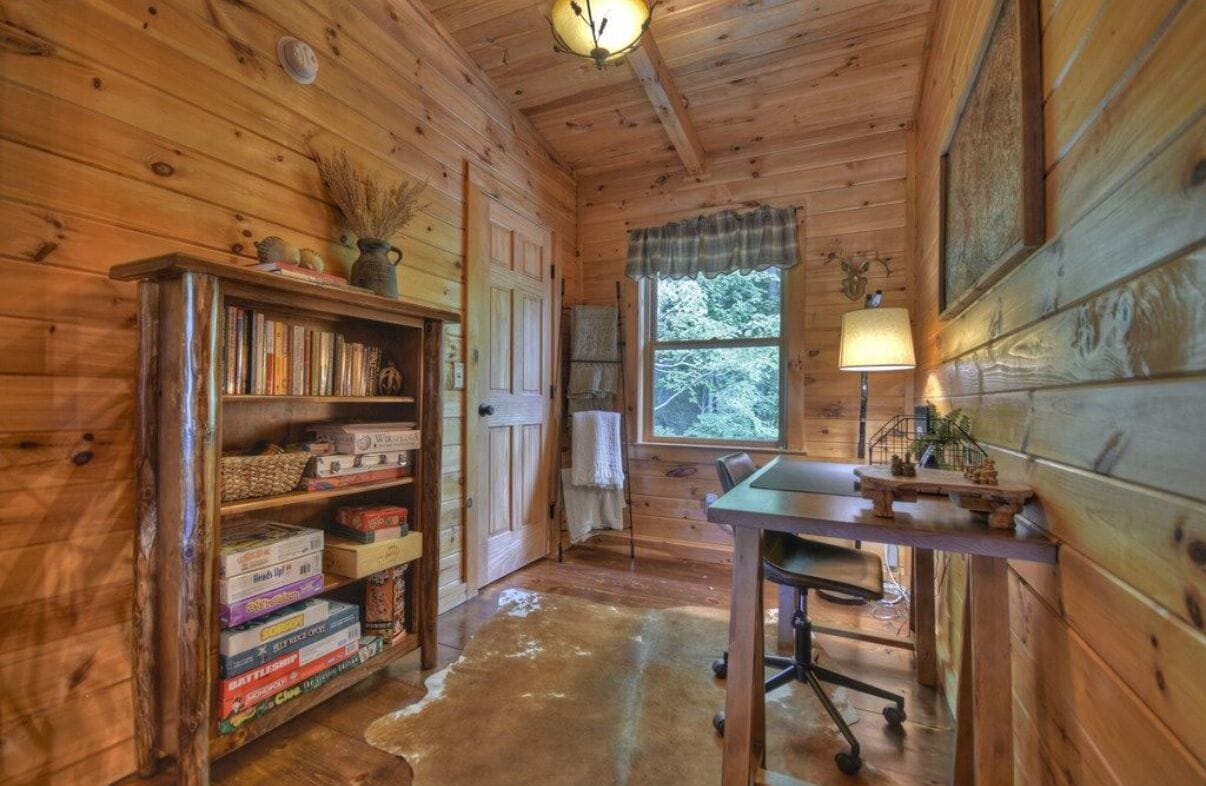 Un accogliente interno di una baita con pareti in legno, una libreria piena di giochi da tavolo, una scrivania con una lampada e una finestra vista alberi.