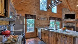 A rustic cabin kitchen with wooden walls, a stone fireplace, and a large window overlooking a deck. - Luxury Pet-Friendly Mountain Cabin - Hot Tub - Views - 20 Min to Blue Ridge (Cherry Log)