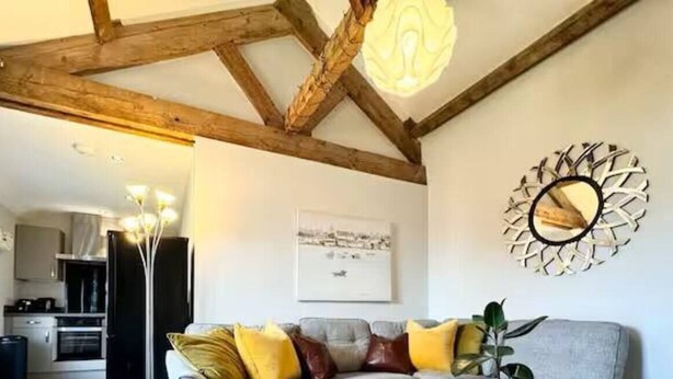 A living room with exposed wooden beams, a grey sofa with yellow and brown cushions, a wall-mounted mirror, and a framed picture. - Warehouse Apartments (Ulverston)
