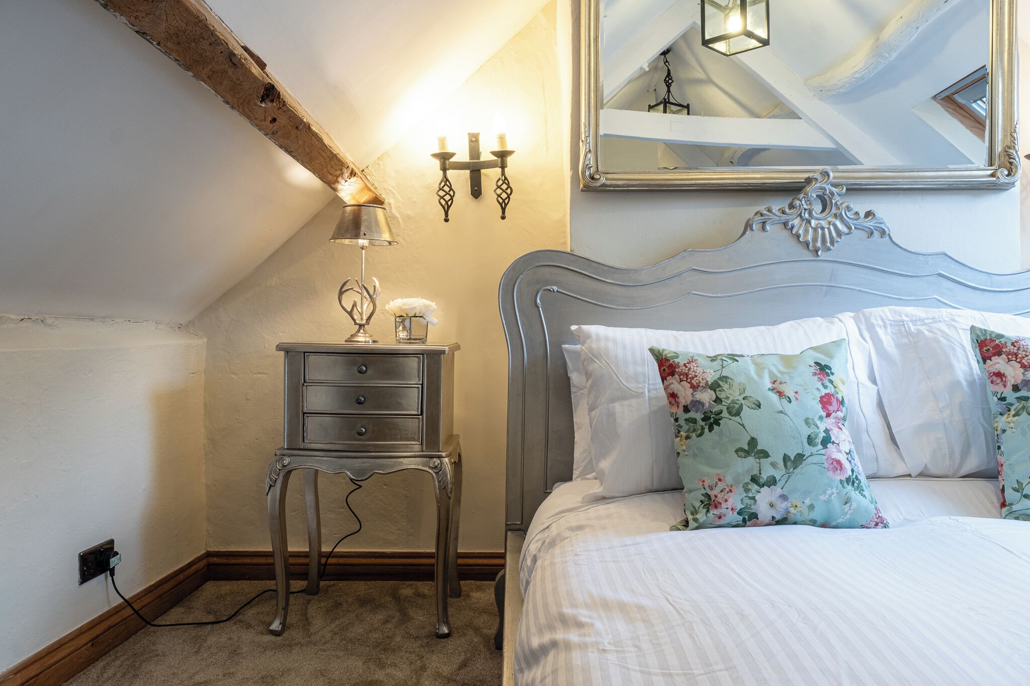 A bedroom with a bed, a nightstand, a lamp, a mirror, and a wall-mounted light fixture.
