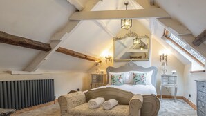 Een slaapkamer met een schuine plafond, een groot bed met bloemenbeddels, een vintage fauteuil, een dressoir en een spiegel met kaarshouders.