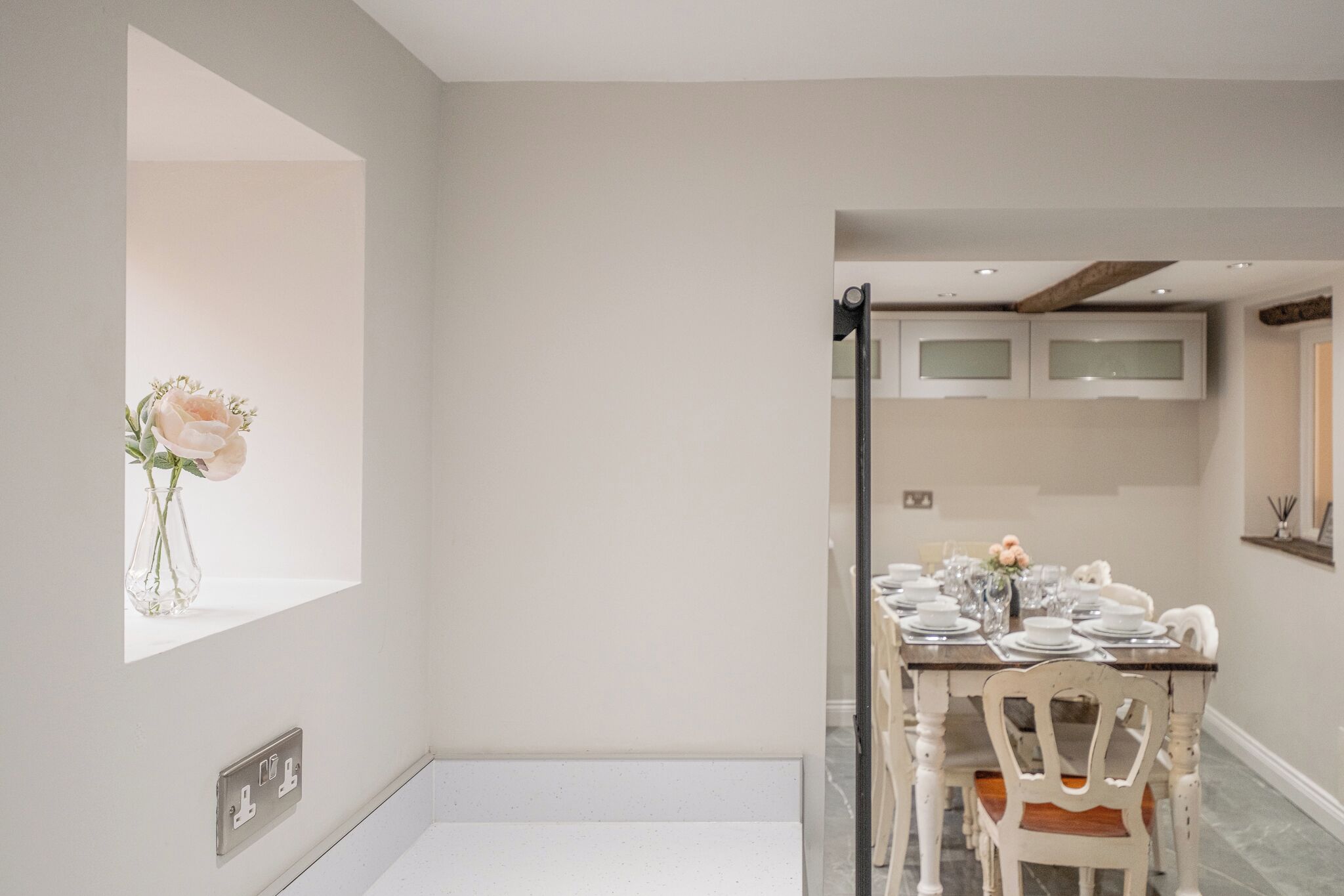 A modern dining area with a white table set for four, a vase with flowers, and a window with a view.