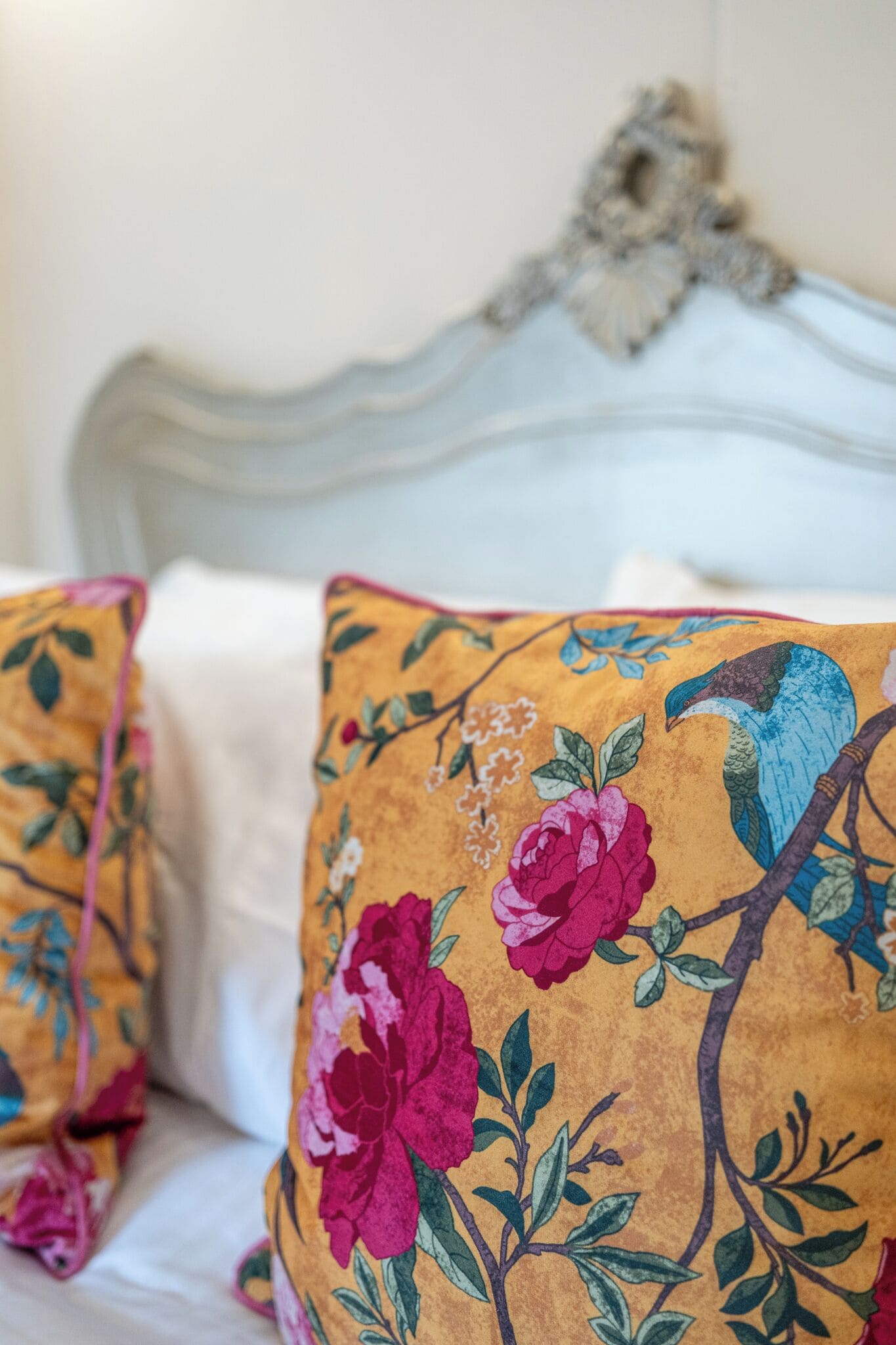 A bed with white bedding and two decorative pillows with floral and bird designs.