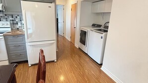 A modern kitchen with a refrigerator, oven, and microwave. There is a dining area with a wooden table and chairs. The floor is wooden, and the walls are white.