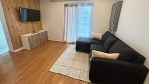 A modern living room with a black sofa, a flat-screen TV mounted on a wooden wall, a small white side table, and a large window with a view of the outdoors.