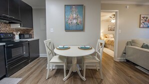 A dining area with a white table and chairs, a kitchen with dark cabinets and a microwave, and a living room with a couch and a painting on the wall. - Tallahassee Gem-Next to Doak and CollegeTown-Ideal for FSU weekend.3 minute walk (Tallahassee)