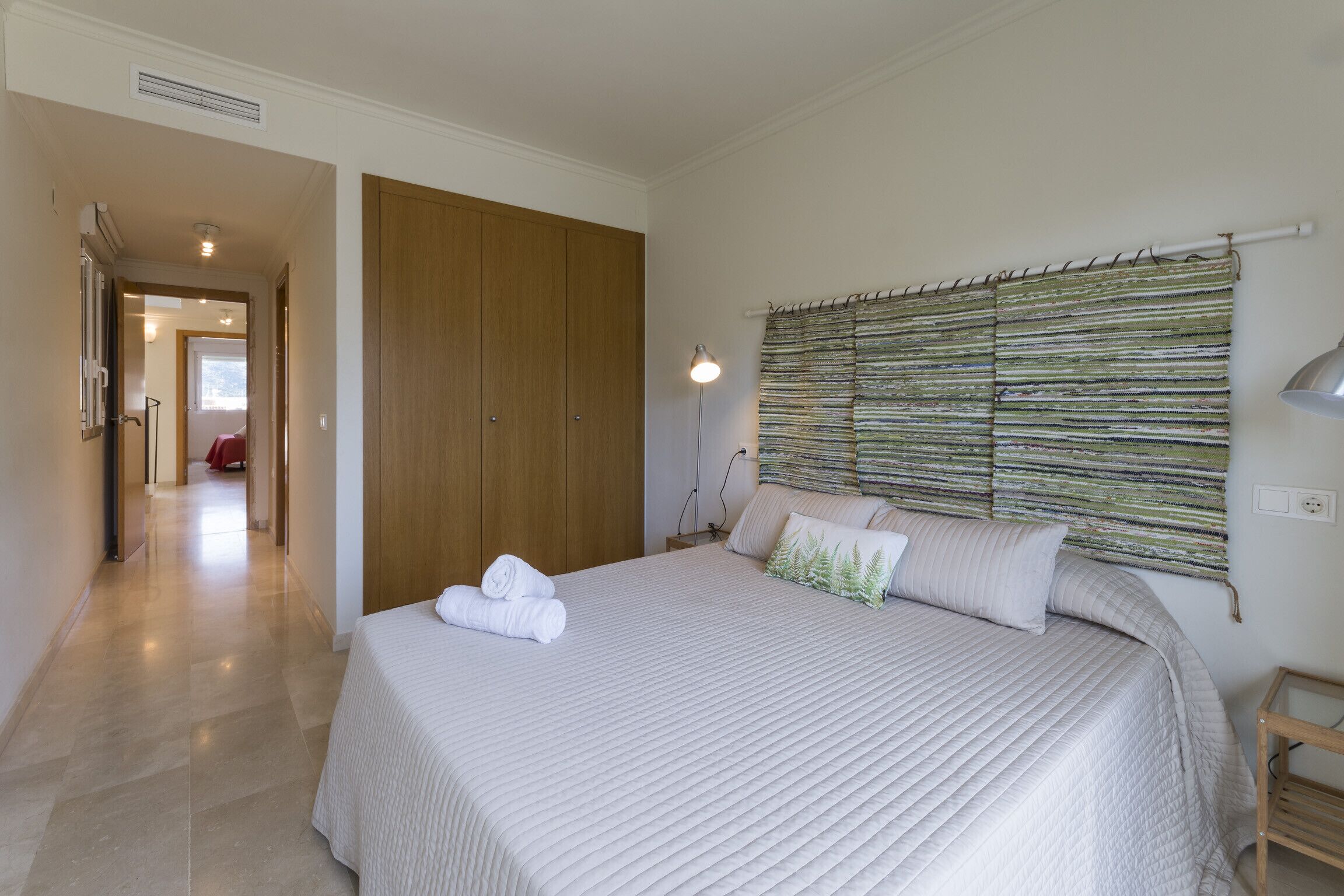 A bedroom with a large bed, a wooden wardrobe, a bedside lamp, and a wall-mounted curtain.