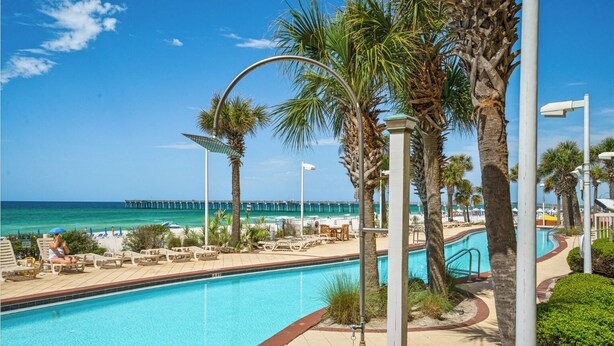 A pool area with lounge chairs, palm trees, and a view of the ocean and pier.