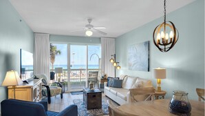 A living room with a view of the ocean, a ceiling fan, a chandelier, and a dining area with a table and chairs.