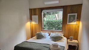 Un letto rifatto con cuscini e coperta, un comodino con lampada e una piccola pianta, e una finestra con vista sul verde.