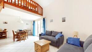 Un vaste espace de vie comprenant un canapé gris, une table basse en bois et une table à manger avec des chaises. Un escalier mène à l’étage supérieur.