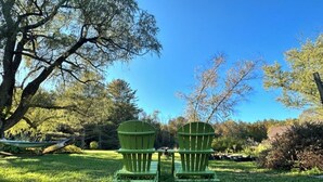 Two green Adirondack chairs in a grassy outdoor area with trees and a clear blue sky.