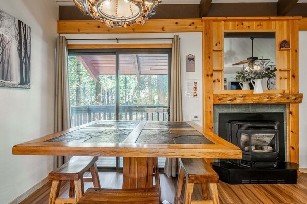 Une table à manger en bois, avec des bancs, une cheminée et une vue sur l’extérieur grâce à une porte vitrée.