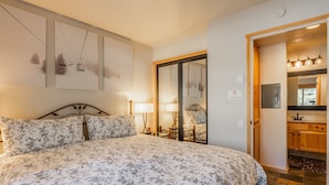 Ein Schlafzimmer mit einem großen Bett, einem Nachttisch, einer Lampe und einem Spiegel. Eine Tür führt zu einem Badezimmer mit Waschbecken und Spiegel.