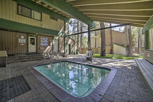 A covered outdoor swimming pool with a brick deck, wooden steps, and a wooden building in the background.