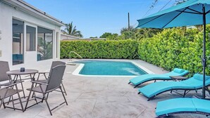 A swimming pool with lounge chairs and a table, surrounded by a hedge and a building. - Coastal Breeze Retreat with Heated Pool (Pembroke Pines)