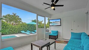 A room with a large window overlooking a swimming pool, a ceiling fan, and a television mounted on the wall. - Coastal Breeze Retreat with Heated Pool (Pembroke Pines)
