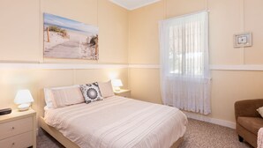 A bedroom with a bed, bedside tables, a nightstand, a window with sheer curtains, a framed picture on the wall, and a brown armchair. - Escape from it all at Crooked Cottage, your peaceful retreat nestled in the heart of Nambucca Heads. (Nambucca Heads)