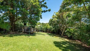 A backyard with a swing set, a wooden deck, and a large tree providing shade. - Escape from it all at Crooked Cottage, your peaceful retreat nestled in the heart of Nambucca Heads. (Nambucca Heads)