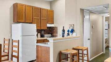 A compact kitchen with wooden cabinets, a white refrigerator, a countertop with a blender, and two wicker stools.