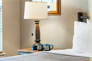 Una cama bien hecha con un almohadón blanco, una mesita de noche de madera con una lámpara, y dos tazas azules con un logotipo.