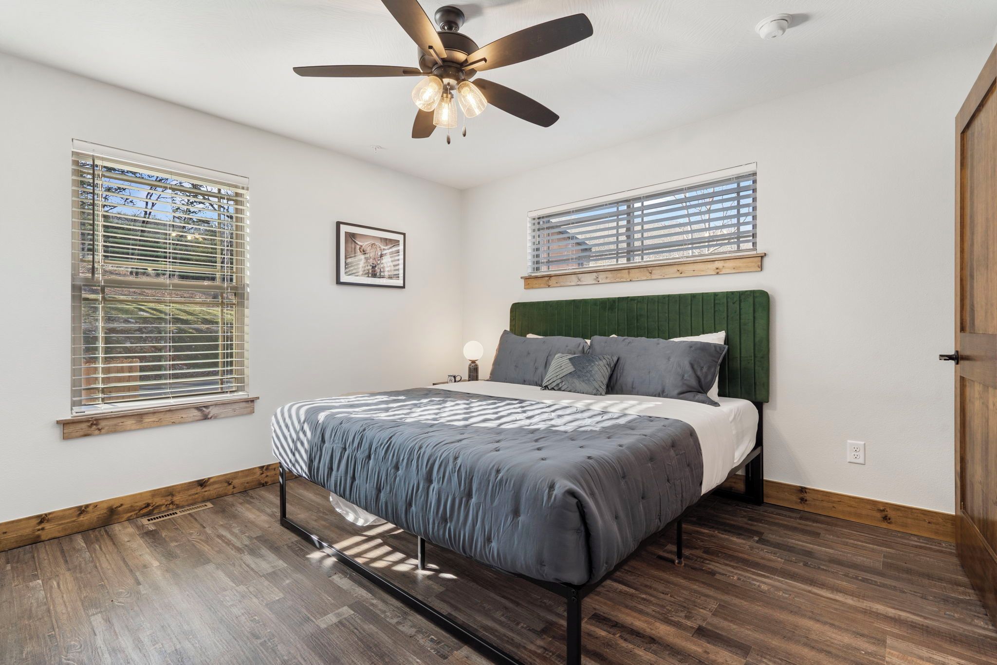 Quarto com cama, ventilador de teto e duas janelas com cortinas.