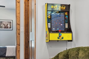Une borne d’arcade Pac-Man fixée à un mur blanc dans une pièce où se trouvent un lit et un tableau encadré.