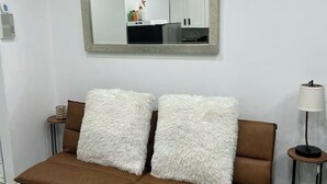A modern living room with a brown sofa, two fluffy white cushions, a large mirror, and a small side table with a lamp. - Studio 2 minute walk to Isla Verde beach (Carolina)