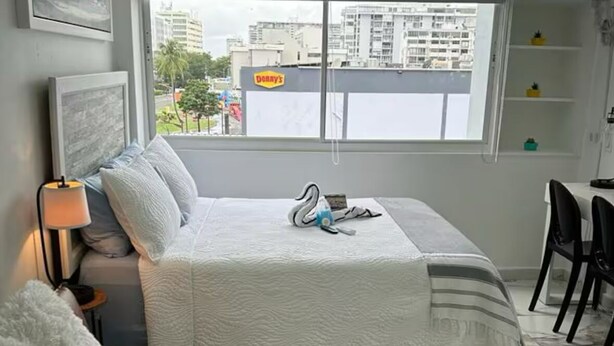A bedroom with a bed, a nightstand, a lamp, a sofa, and a window with a city view. - Studio 2 minute walk to Isla Verde beach (Carolina)