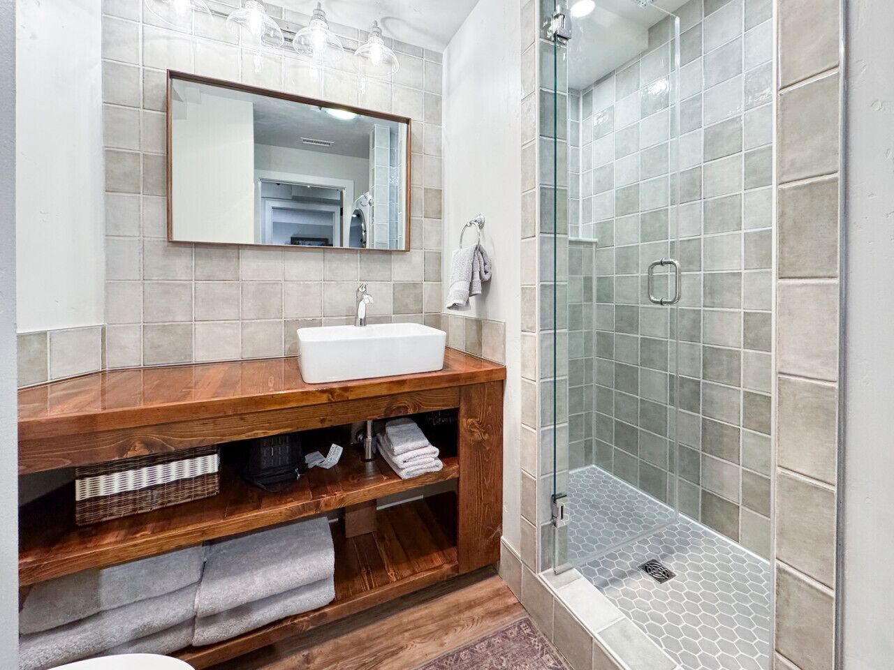 Un baño con tocador de madera, lavamanos blanco, espejo y cabina de ducha de vidrio.