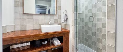 Un baño con tocador de madera, lavamanos blanco, espejo y cabina de ducha de vidrio.