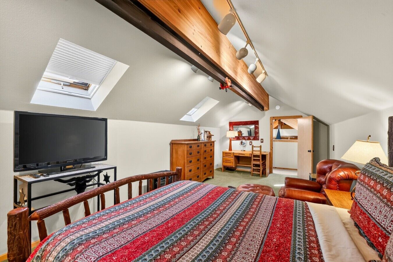 A bedroom with a large bed, a desk, a dresser, a TV, and a skylight.