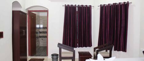 A hotel room with a bed, two chairs, a small table, a trash bin, and a shower area.