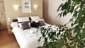 Un lit bien fait, avec une literie blanche et grise, une tête de lit agrémentée de lampes murales et une plante au premier plan.