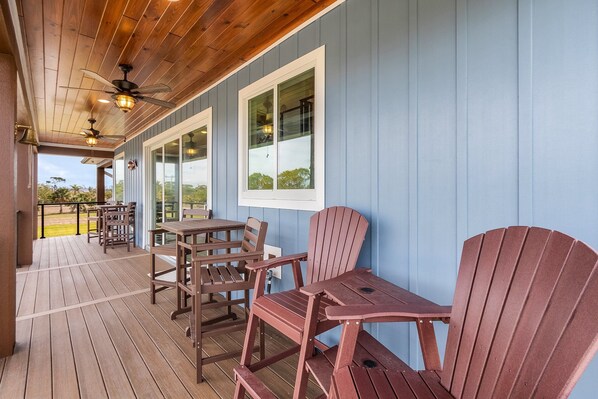 A covered outdoor seating area with wooden tables and chairs, a ceiling fan, and large windows.