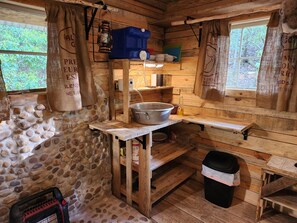 A rustic cabin interior with a stone wall, wooden shelves, a metal basin, and a black trash bin. - Raelean - A unique off-grid tiny cabin (Anniston)