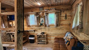 L’intérieur d’un chalet rustique, avec un plancher en bois, un canapé marron, une petite table, une lampe et une fenêtre avec des rideaux.