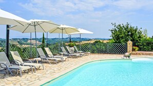 Un área junto a la piscina con sillones de descanso y sombrillas, una piscina con baldosas de piedra y vistas al campo.