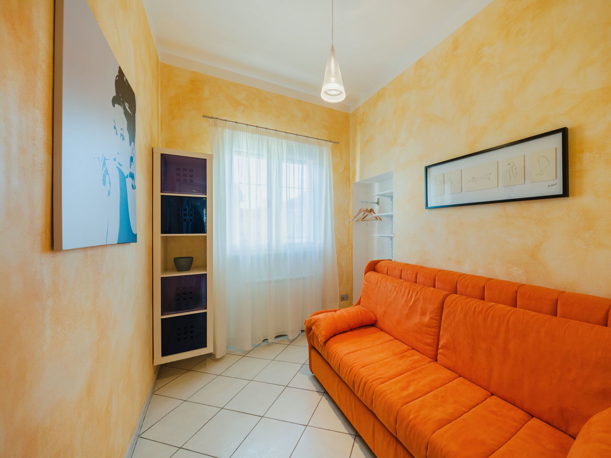 A room with a tiled floor, a white wardrobe, a framed artwork, and an orange sofa.