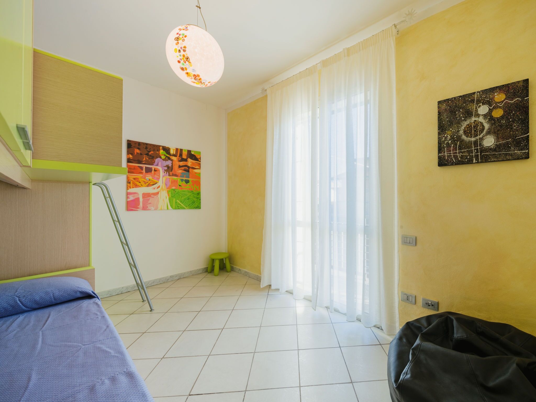 A room with a bed, a green chair, a small stool, a painting, and a window with curtains.