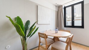Un pequeño comedor con una mesa redonda y cuatro sillas, una planta en maceta, una obra de arte enmarcada y una ventana con cortinas.